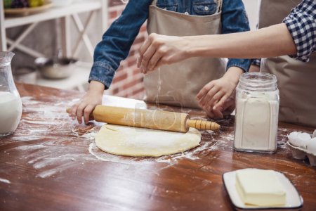 Téléchargez les photos : Image recadrée de la petite fille et de sa mère dans des tabliers aplatissant la pâte à l'aide d'un rouleau à pâtisserie dans la cuisine - en image libre de droit