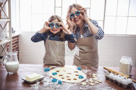 Photo for Cute little girl and her beautiful mom in aprons are smiling while preparing cookies using cookie cutters in the kitchen - Royalty Free Image