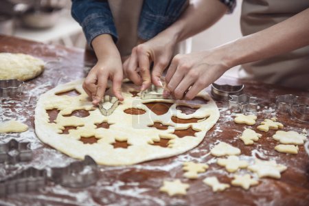 Téléchargez les photos : Image recadrée de la petite fille et de sa mère dans des tabliers préparant des biscuits à l'aide de coupe-biscuits dans la cuisine - en image libre de droit