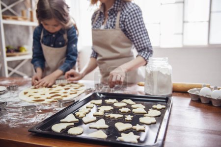 Foto de Linda niña y su hermosa mamá en delantales están preparando galletas utilizando cortadores de galletas en la cocina - Imagen libre de derechos