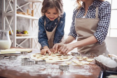 Foto de Linda niña y su hermosa mamá en delantales sonríen mientras preparan galletas usando cortadores de galletas en la cocina - Imagen libre de derechos