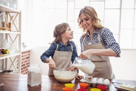 Foto de Linda niña y su hermosa mamá en delantales sonríen mientras preparan masa para magdalenas en la cocina - Imagen libre de derechos