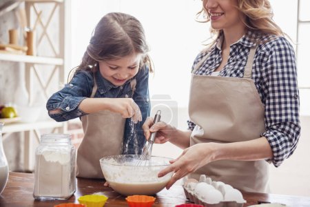 Foto de Linda niña y su hermosa mamá en delantales sonríen mientras preparan masa para magdalenas en la cocina - Imagen libre de derechos