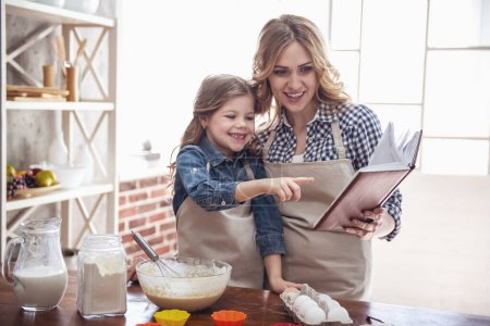Foto de Linda niña y su hermosa mamá en delantales están leyendo receta y sonriendo mientras amasa la masa en la cocina - Imagen libre de derechos