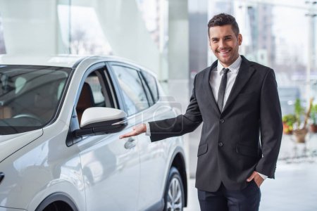 Photo for Handsome businessman in suit is looking at camera and smiling while standing near the car in car dealership - Royalty Free Image