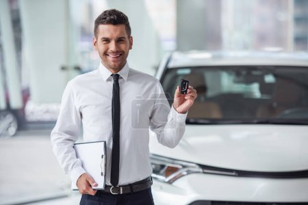 Foto de Hermoso concesionario de automóviles trabajador de traje está sosteniendo la llave, mirando a la cámara y sonriendo mientras está de pie cerca del coche - Imagen libre de derechos