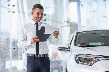 Photo for Handsome car dealership worker in suit is holding a folder and smiling while standing near the car - Royalty Free Image
