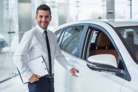 Foto de Apuesto concesionario de automóviles en traje está sosteniendo una carpeta, mirando a la cámara y sonriendo mientras está de pie cerca del coche - Imagen libre de derechos