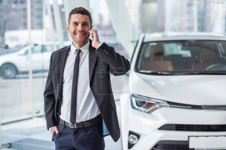 Foto de Hombre de negocios guapo en traje está hablando en el teléfono móvil y sonriendo mientras está de pie cerca del coche en concesionario de automóviles - Imagen libre de derechos