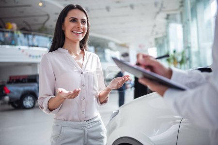 Foto de Hermosa mujer joven está hablando con el trabajador de concesionario de automóviles guapo al elegir un coche en concesionario - Imagen libre de derechos