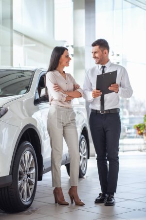 Foto de Hermosa mujer joven está hablando con el trabajador de concesionario de automóviles guapo al elegir un coche en concesionario - Imagen libre de derechos