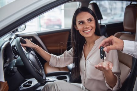 Photo for Beautiful young woman is getting key and smiling while sitting in a new car in dealership - Royalty Free Image