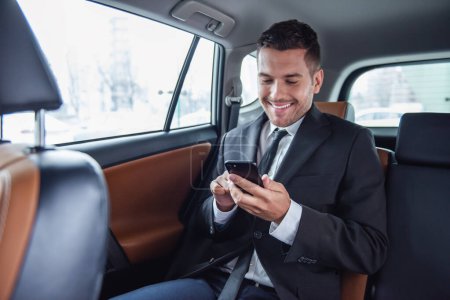 Foto de Hombre de negocios guapo en traje está utilizando un teléfono inteligente y sonriendo mientras está sentado en el asiento trasero en el coche - Imagen libre de derechos