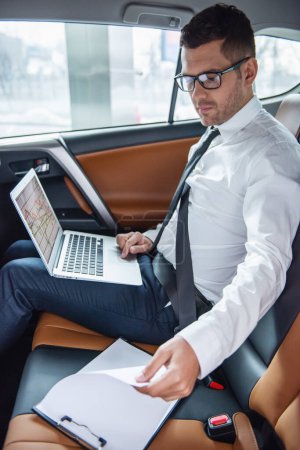 Téléchargez les photos : Beau homme d'affaires en costume et lunettes utilise un ordinateur portable et d'étudier les documents tout en conduisant sur le siège arrière dans la voiture - en image libre de droit