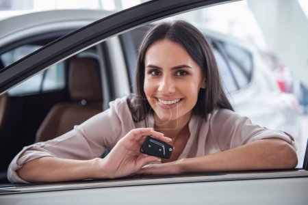 Foto de Visitando concesionario de coches. Hermosa joven mujer está mostrando la llave de su nuevo coche, mirando a la cámara y sonriendo - Imagen libre de derechos