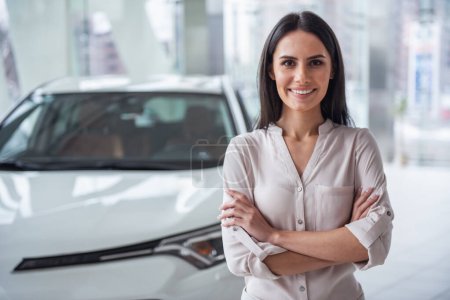 Photo for Visiting car dealership. Beautiful young woman is looking at camera and smiling while standing with crossed arms near her new car - Royalty Free Image