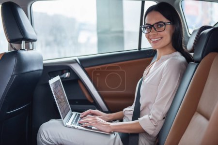 Téléchargez les photos : Belle femme d'affaires dans les lunettes utilise un ordinateur portable et souriant tout en étant assis sur le siège arrière dans la voiture - en image libre de droit