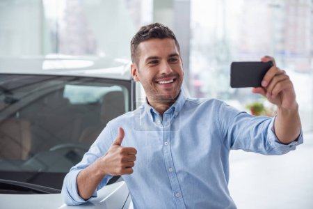 Foto de Visitando concesionario de coches. Hombre guapo está haciendo selfie con su nuevo coche, mostrando el pulgar y sonriendo - Imagen libre de derechos