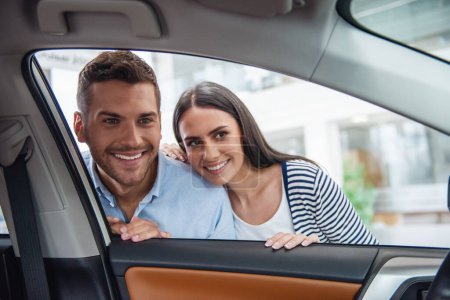 Foto de Visitando concesionario de coches. Hermosa pareja está examinando un coche nuevo y sonriendo - Imagen libre de derechos