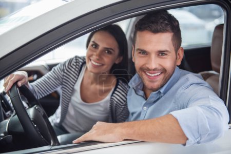 Foto de Visitando concesionario de coches. Hermosa pareja está mirando a la cámara y sonriendo mientras está sentado en su nuevo coche - Imagen libre de derechos