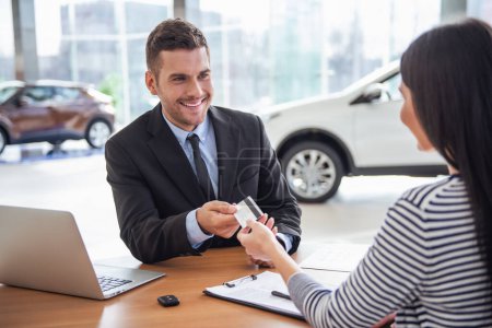 Photo for Visiting car dealership. Handsome sales manager is taking a credit card from the client and smiling - Royalty Free Image