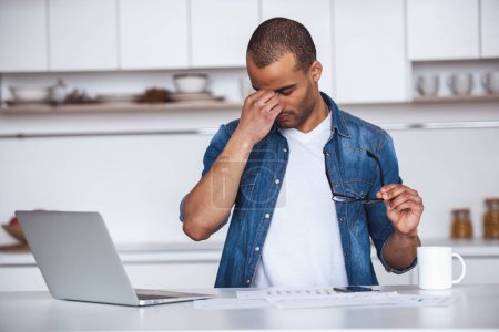 Foto de Hombre de negocios cansado guapo está masajeando puente nasal mientras trabaja con un ordenador portátil en la cocina - Imagen libre de derechos