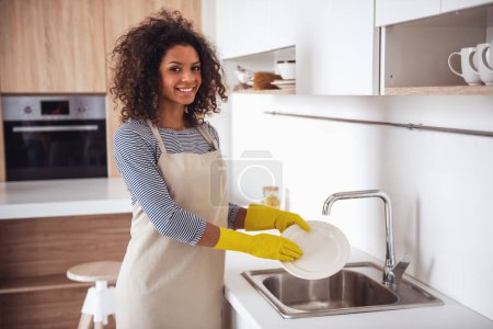 Téléchargez les photos : Belle jeune femme regarde la caméra et sourit tout en lavant la vaisselle dans la cuisine - en image libre de droit