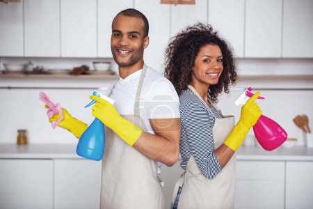 Foto de Hermosa pareja joven está sosteniendo detergentes, mirando a la cámara y sonriendo mientras limpia la cocina - Imagen libre de derechos