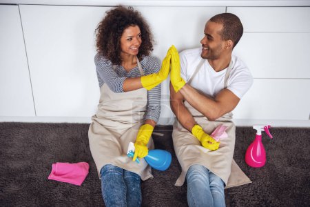Photo for Beautiful young couple is giving high five and smiling while sitting on the floor in kitchen after cleaning it - Royalty Free Image