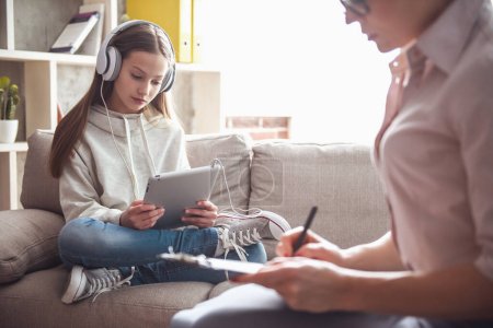 Photo for Teenage girl at the psychotherapist. Doctor is making notes while girl is sitting in headphones on sofa - Royalty Free Image