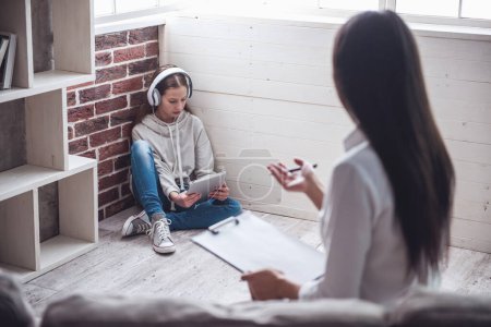 Photo for Teenage girl at the psychotherapist. Doctor is making notes while girl is sitting in headphones in the corner on the floor - Royalty Free Image