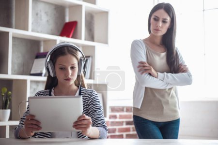 Foto de Adolescente con problemas en los auriculares está utilizando una tableta digital mientras su madre está de pie con los brazos cruzados en el fondo - Imagen libre de derechos