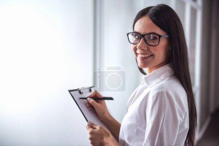 Foto de Hermoso joven psicoterapeuta está tomando notas, mirando a la cámara y sonriendo mientras está de pie en la oficina - Imagen libre de derechos