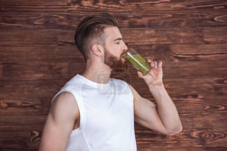 Photo for Handsome young bearded sportsman is drinking nutritious cocktail, on wooden background - Royalty Free Image