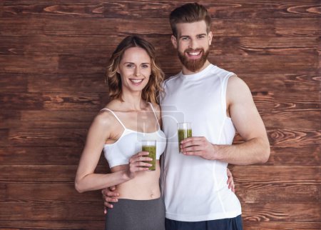 Photo for Beautiful young sports people are holding nutritious cocktails, looking at camera and smiling, on wooden background - Royalty Free Image