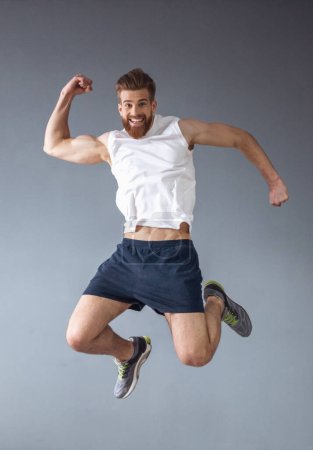 Photo for Handsome young bearded sportsman is showing muscles, jumping and smiling, on gray background - Royalty Free Image
