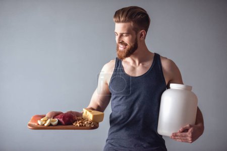 Photo for Handsome young bearded sportsman is holding a bottle with sport nutrition and a wooden tray with healthy food, on gray background - Royalty Free Image