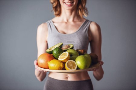 Photo for Cropped image of beautiful young sportswoman holding a tray with fruits and smiling, on gray background - Royalty Free Image
