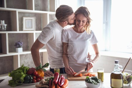 Foto de Hermosa pareja joven está hablando y sonriendo mientras cocina alimentos saludables en la cocina en casa. El hombre está besando a su novia en la mejilla - Imagen libre de derechos