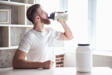 Photo for Handsome young bearded sportsman is drinking while preparing sport nutrition in kitchen at home - Royalty Free Image