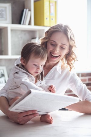 Foto de Hermosa mamá de negocios en traje está leyendo un periódico con su lindo bebé y sonriendo, en casa - Imagen libre de derechos