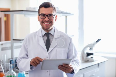 Téléchargez les photos : Médecin beau en manteau blanc utilise une tablette numérique, regardant la caméra et souriant tout en se tenant debout en laboratoire - en image libre de droit