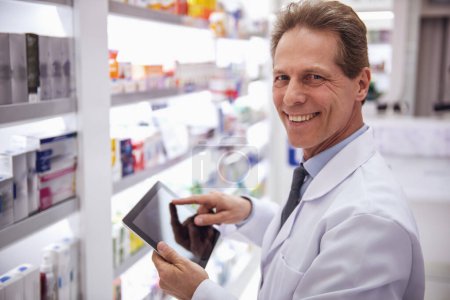 Téléchargez les photos : Beau pharmacien utilise un comprimé numérique et souriant tout en vérifiant les médicaments en pharmacie - en image libre de droit