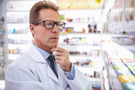 Photo for Handsome pharmacist in eyeglasses is rubbing his chin while searching for drug in pharmacy - Royalty Free Image