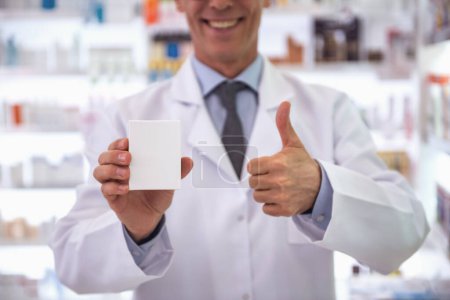 Téléchargez les photos : Image recadrée d'un beau pharmacien tenant un médicament, montrant le signe Ok et souriant alors qu'il se tenait debout en pharmacie - en image libre de droit