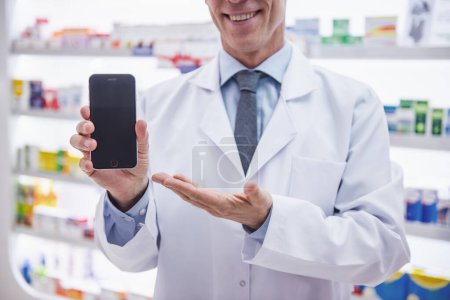Photo for Cropped image of handsome pharmacist holding a smart phone and smiling while standing in pharmacy - Royalty Free Image