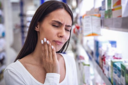 Photo for Beautiful young woman is having a toothache while choosing a remedy at the pharmacy - Royalty Free Image
