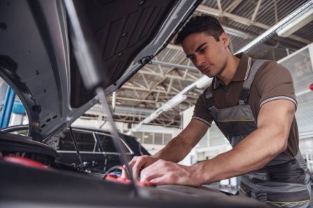 Photo for Handsome young auto mechanic in uniform is repairing car in auto service - Royalty Free Image