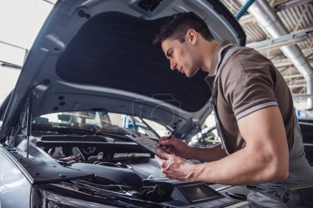 Photo for Handsome young auto mechanic in uniform is making notes while examining car in auto service - Royalty Free Image
