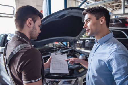 Photo for At the auto service. Handsome young auto mechanic in uniform is talking with a client and showing a document to fill - Royalty Free Image